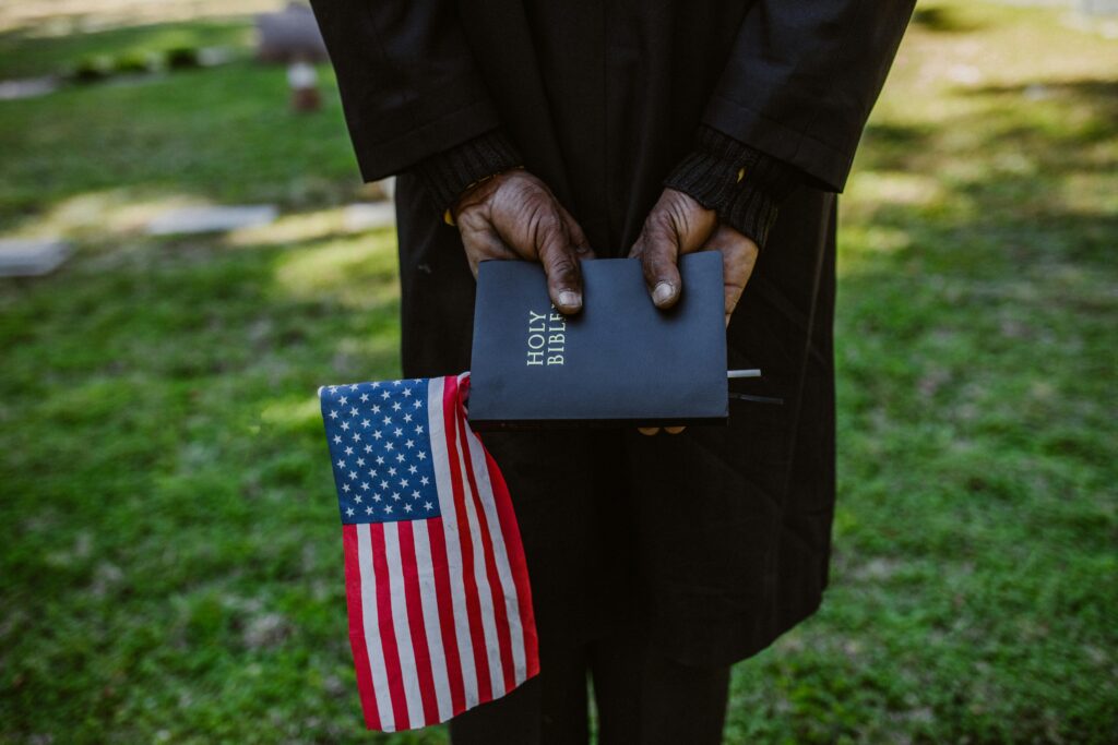 Close-up of hands holding a Holy Bible and American flag in a green outdoor setting.